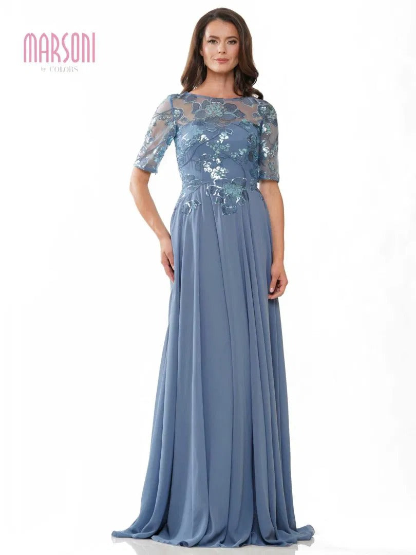 A woman models the Marsoni M286 long formal dress in slate blue, featuring sheer floral lace on the bodice and sleeves, while standing against a plain white background.